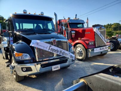 Photo shows the fronts of two semitrucks. Each have banners hung across the front: one says "In Memory of Francis Wallace Foster" and the other has two banners that say "In Memory of Ginny Elder" and "In Memory of Buddy Elder."