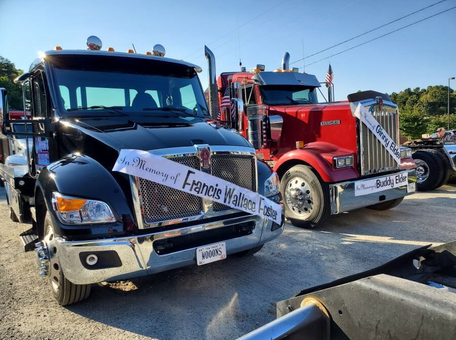 Photo shows the fronts of two semitrucks. Each have banners hung across the front: one says "In Memory of Francis Wallace Foster" and the other has two banners that say "In Memory of Ginny Elder" and "In Memory of Buddy Elder."