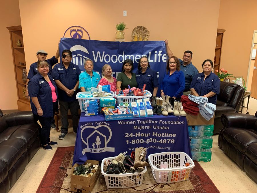 A group of 10 people stand with donations for Mujeres Unidas. Among the donations are water, shoes, towels, and toothbrushes. Behind them is a WoodmenLife banner. On the table in front of the people is a banner for Mujeres Unidas / Women Together Foundation.