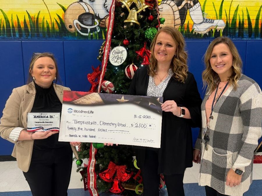 Three women are holding an oversized WoodmenLife-branded check. The check is made out to Tompkinsville Elementary School in the amount of $2,500. The woman on the left is holding a small plaque that says "America Is ..." at the top. Behind the group is a Christmas tree.