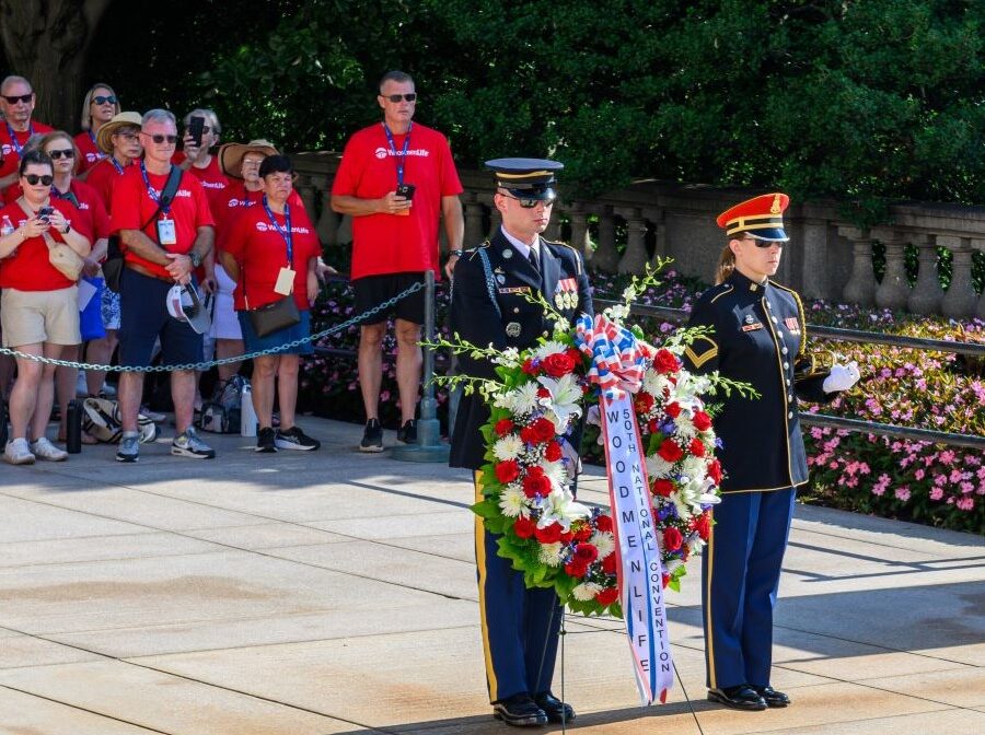 Photo shows two members of the military standing with a large wreath. The wreath is made up of red, white, and blue flowers, and has a red, white, and blue ribbon that says "WoodmenLife 50th National Convention." Behind the service members and the wreath is a crowd of people all wearing red WoodmenLife-branded T-shirts.