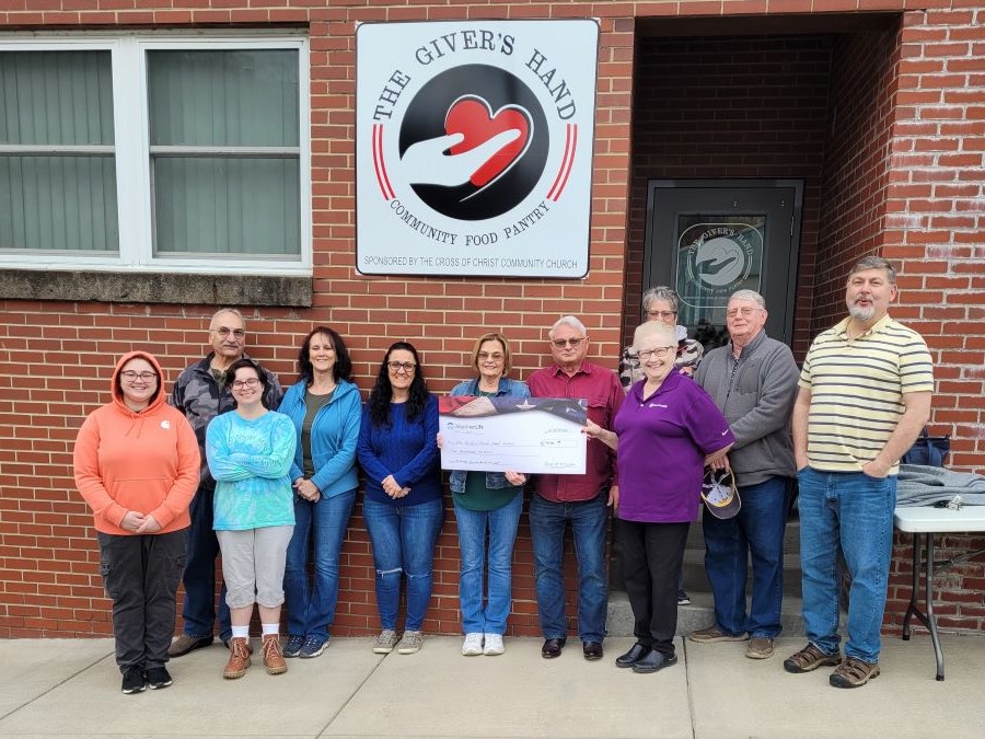 11 people stand together holding a WoodmenLife-branded oversized check. They are standing outside a building; on the building is a sign for The Giver's Hand Community Food Pantry.