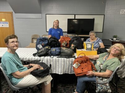 Four women stand and sit around a table filled with backpacks and school supplies.