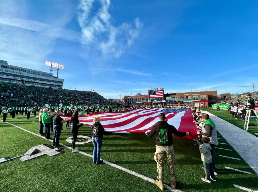 Dozens of volunteers hold a 30-foot-by-60-foot American flag. They are displaying the flag in the middle of a football field.