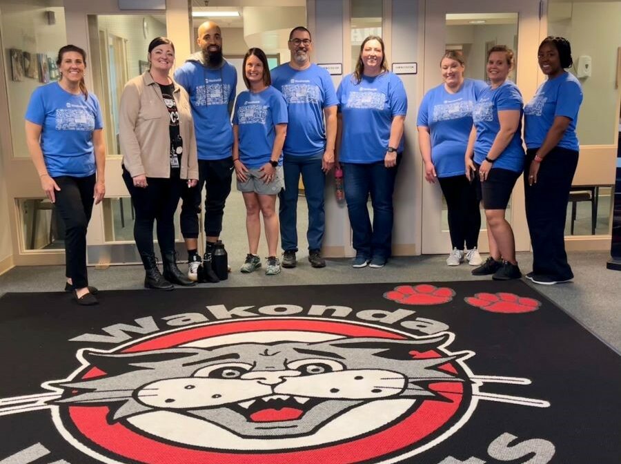 Nine people stand together, eight of whom are wearing blue WoodmenLife-branded t-shirts. They are standing around a floor mat that says "Wakonda" and has the school's mascot, a wildcat.