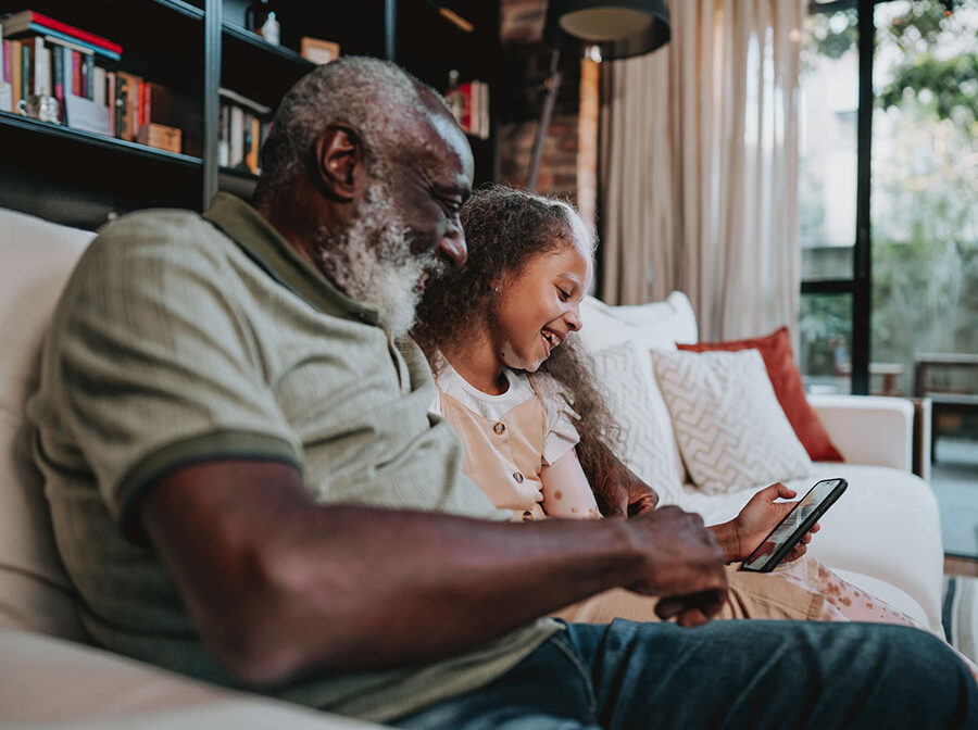 Stock photo shows an older man sitting on a couch with a young girl — they appear to be a grandfather and granddaughter. They are both looking at a cellphone and smiling.