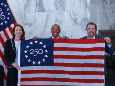 Photo shows three people holding up a commemorative Betsy Ross 250 U.S. flag. On the flag, in the upper-left blue square, is the number 250 with 13 stars around it.