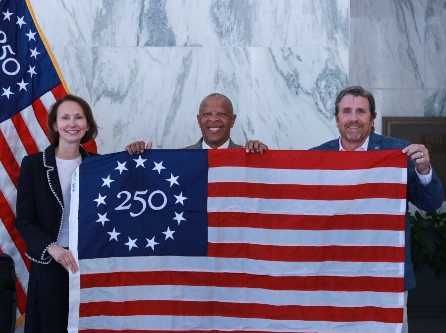 Photo shows three people holding up a commemorative Betsy Ross 250 U.S. flag. On the flag, in the upper-left blue square, is the number 250 with 13 stars around it.