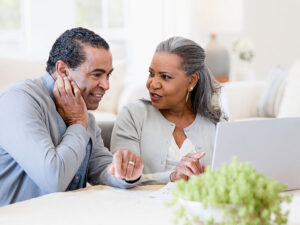 Stock photo shows an older man and woman sitting at a table. They are talking to one another as they look over an open laptop in front of them.