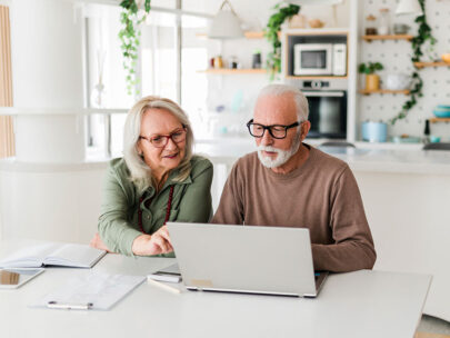 Stock photo shows an elderly man and woman sitting at a table. On the table in front of them is an open laptop, a calculator, and various papers. Behind them is a kitchen.