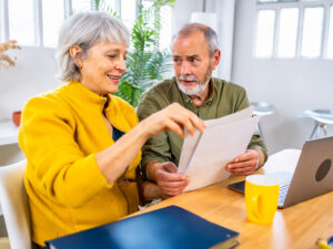 Stock photo shows an older couple sitting at a table. There is an open laptop and a coffee cup on the table in front of them. They are holding paperwork that they're looking over.