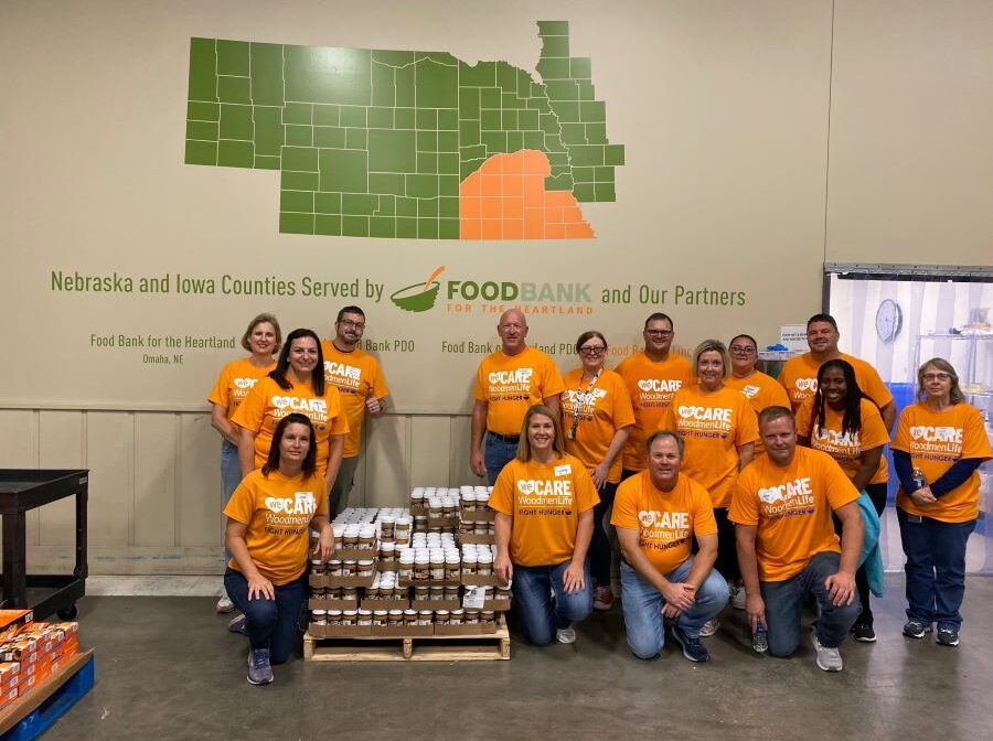 A group of WoodmenLife associates, all wearing orange t-shirts, poses against a wall that shows a map of the Nebraska and Iowa counties served by the Food Bank for the Heartland. WoodmenLife emphasizes the importance of giving back to the community.