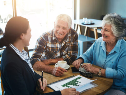 Stock photo shows an older couple sitting at a table with a financial professional. On the table in front of them are coffee cups. The professional is holding a paper with a graph on it in one hand and a pen in the other. Everyone is smiling.