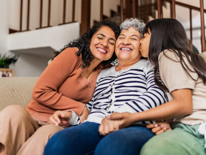 Stock photo shows three people sitting together on a couch. In the center is an older woman. On one side of her is a middle-age woman, and on the other side of her is a young girl; they are both cuddled in close and embracing the older woman in the middle. All three people are smiling.
