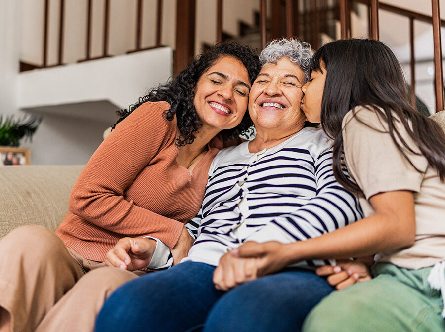 Stock photo shows three people sitting together on a couch. In the center is an older woman. On one side of her is a middle-age woman, and on the other side of her is a young girl; they are both cuddled in close and embracing the older woman in the middle. All three people are smiling.