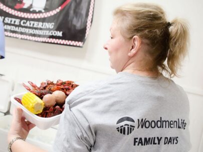 Photo shows a woman whose back is toward the camera and she is turning to look over her shoulder. The back of her T-shirt says "WoodmenLife Family Days." She is holding a plate with crawfish, potatoes, and corn-on-the-cob.