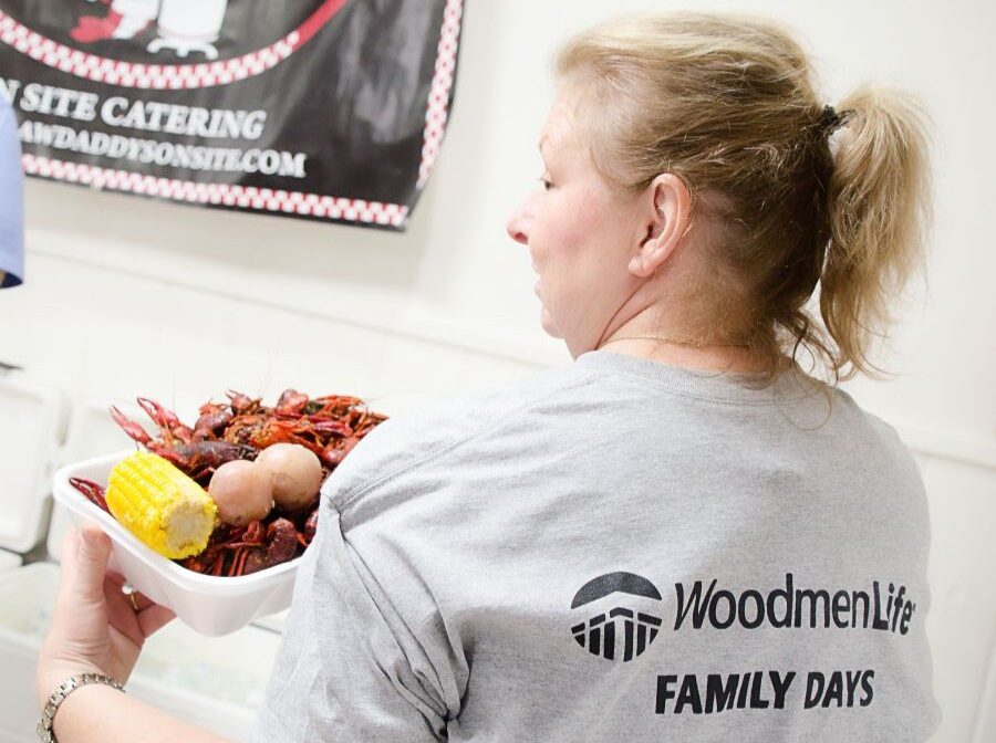Photo shows a woman whose back is toward the camera and she is turning to look over her shoulder. The back of her T-shirt says "WoodmenLife Family Days." She is holding a plate with crawfish, potatoes, and corn-on-the-cob.