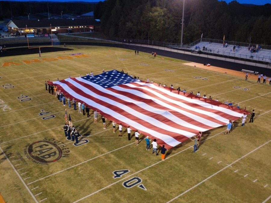 Photo is an overhead view of an oversized American flag being displayed in the center of a football field. Volunteers are standing around the edges of the flag, holding it.