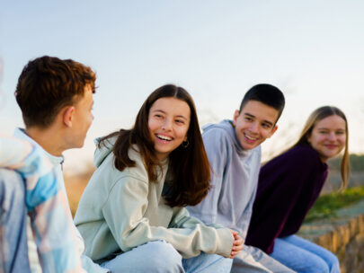 Stock photo shows four teenagers sitting outside. They are looking at one another and smiling. There is blue sky behind them.
