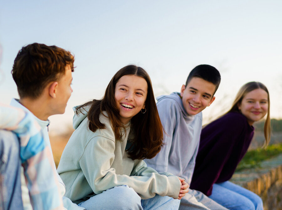 Stock photo shows four teenagers sitting outside. They are looking at one another and smiling. There is blue sky behind them.